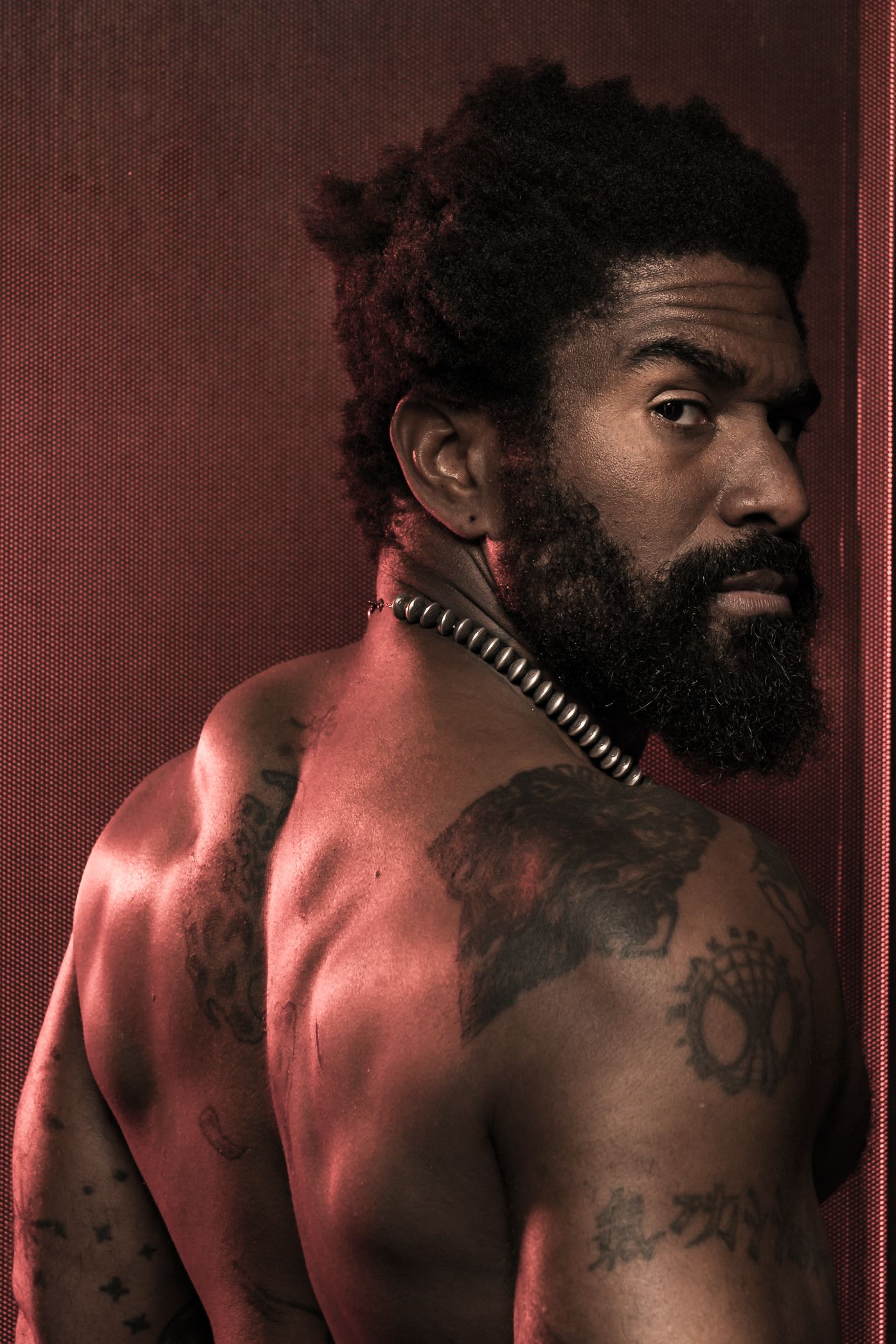 Dramatic artistic portrait with red lighting shows shirtless man with beard and tribal necklace displaying shoulder tattoo.