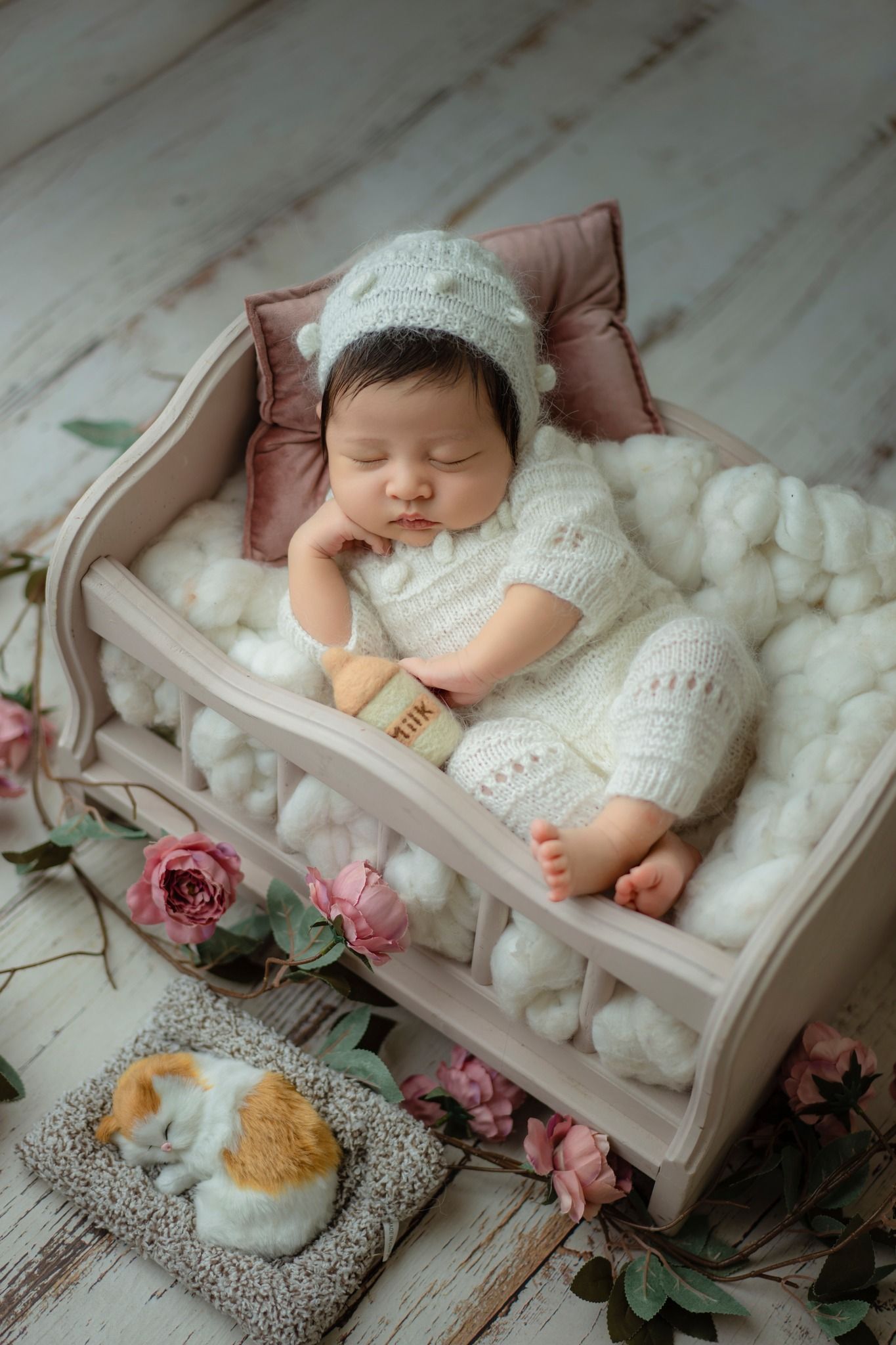 Sleeping baby in knitted white outfit rests in decorative bed with roses and stuffed bunny.