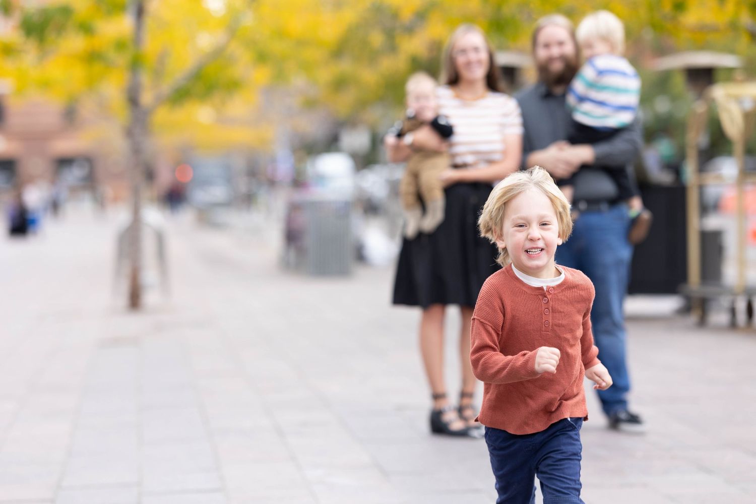 A child runs joyfully down a sidewalk during autumn with people in the background.
