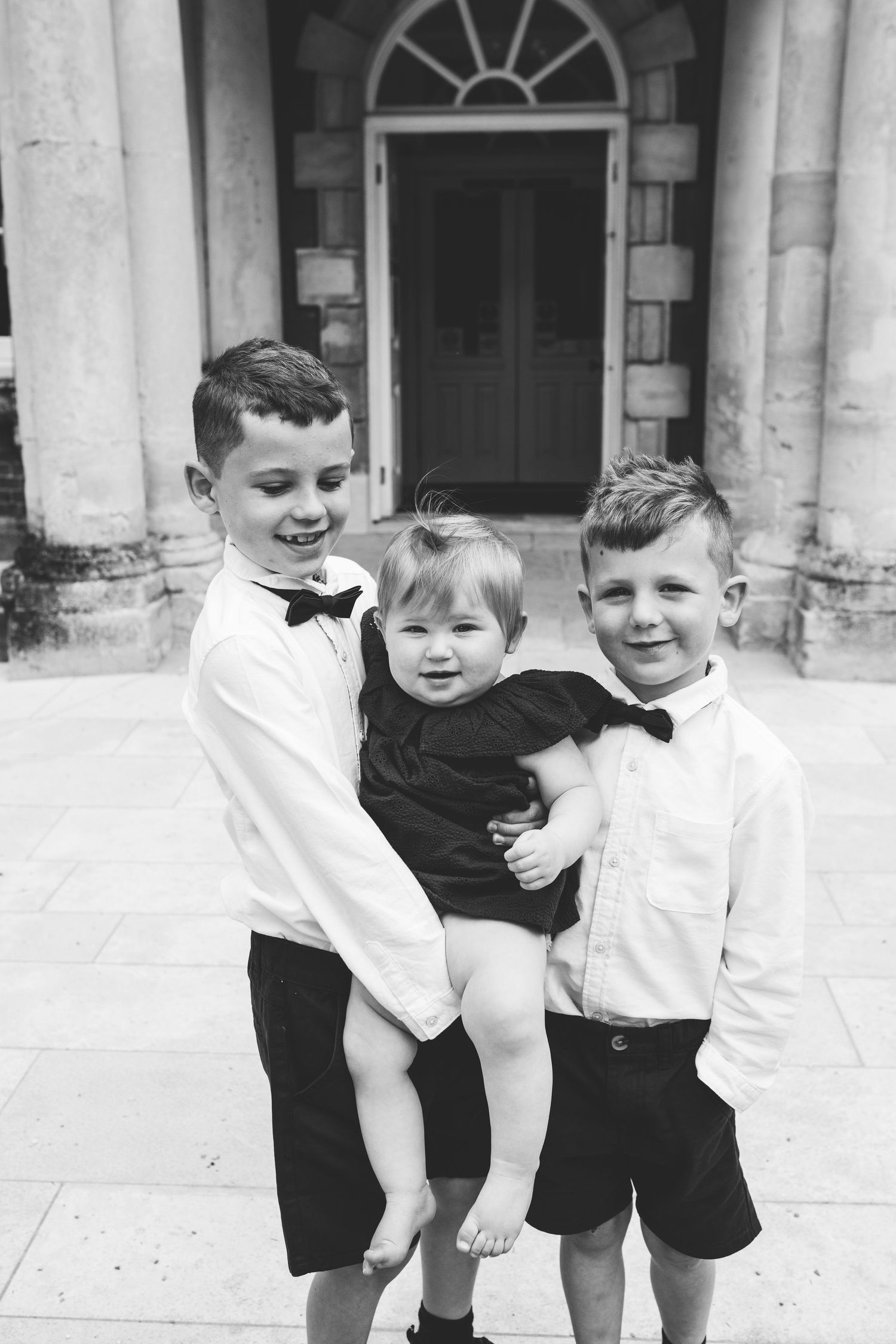 Three young children in white shirts and black shorts play together on a stone porch.
