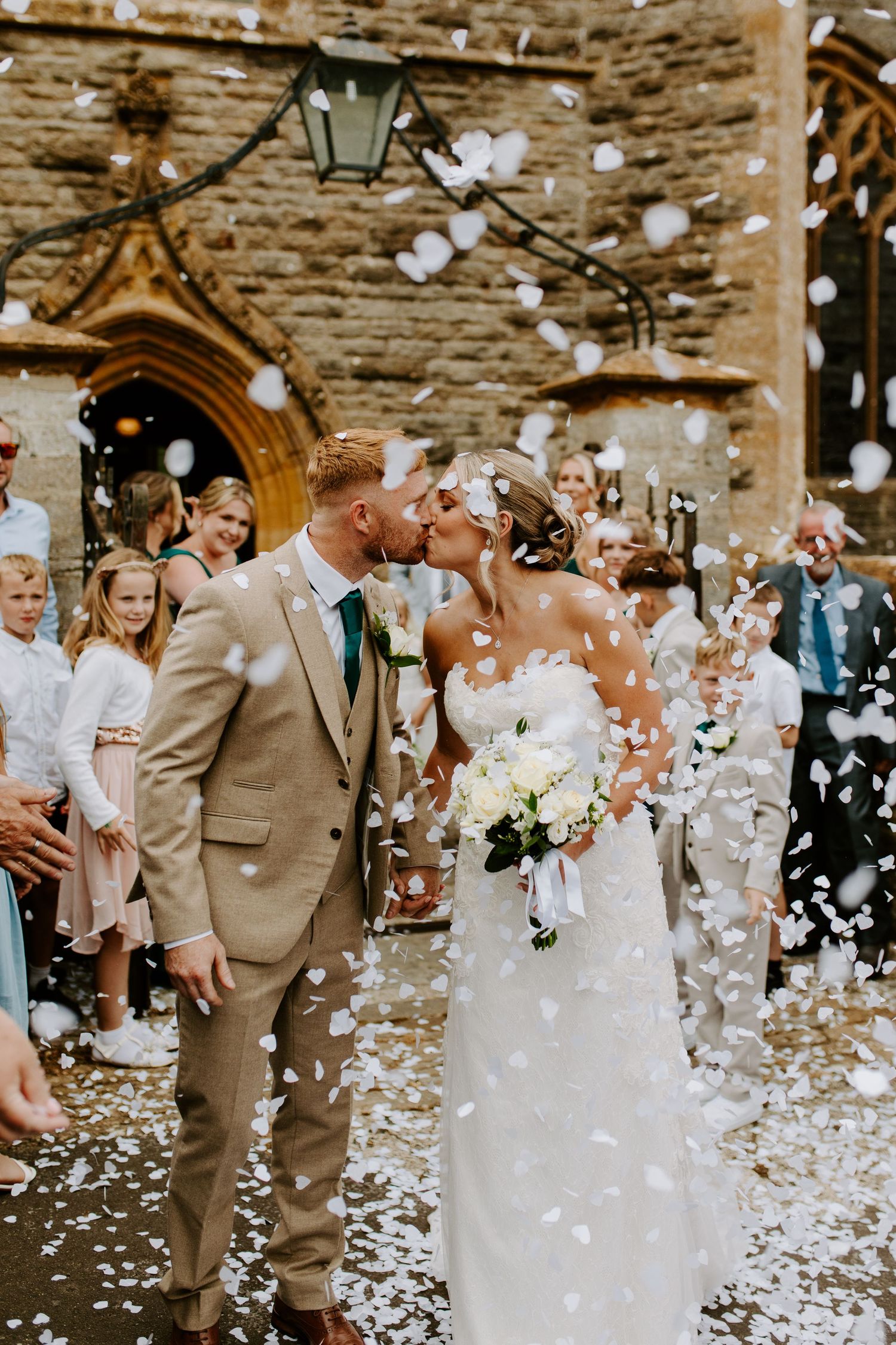 Wedding celebration outside stone church with white flower petals being thrown in celebration.