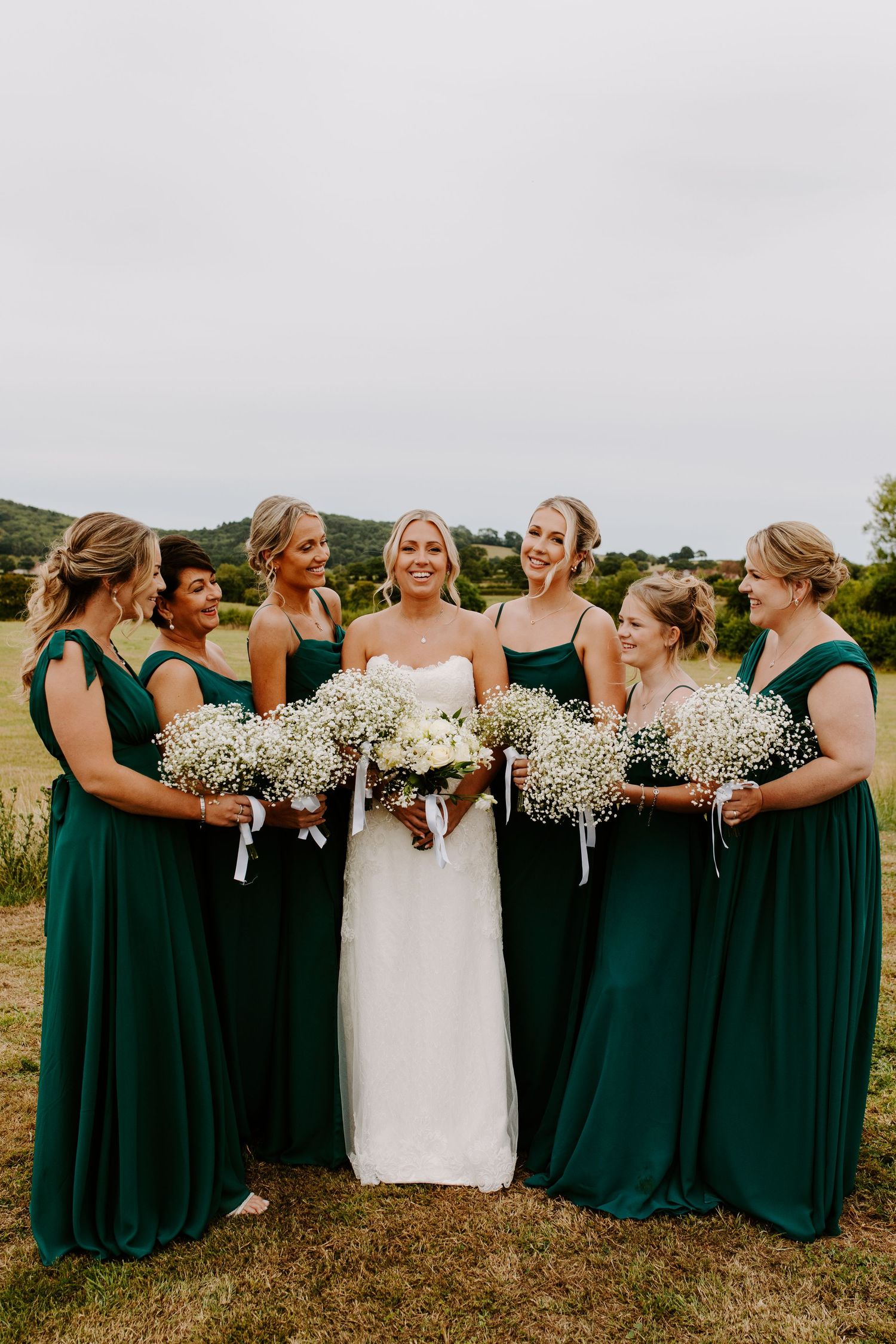 Wedding party in emerald green bridesmaid dresses and white gown holding baby's breath bouquets in outdoor setting.