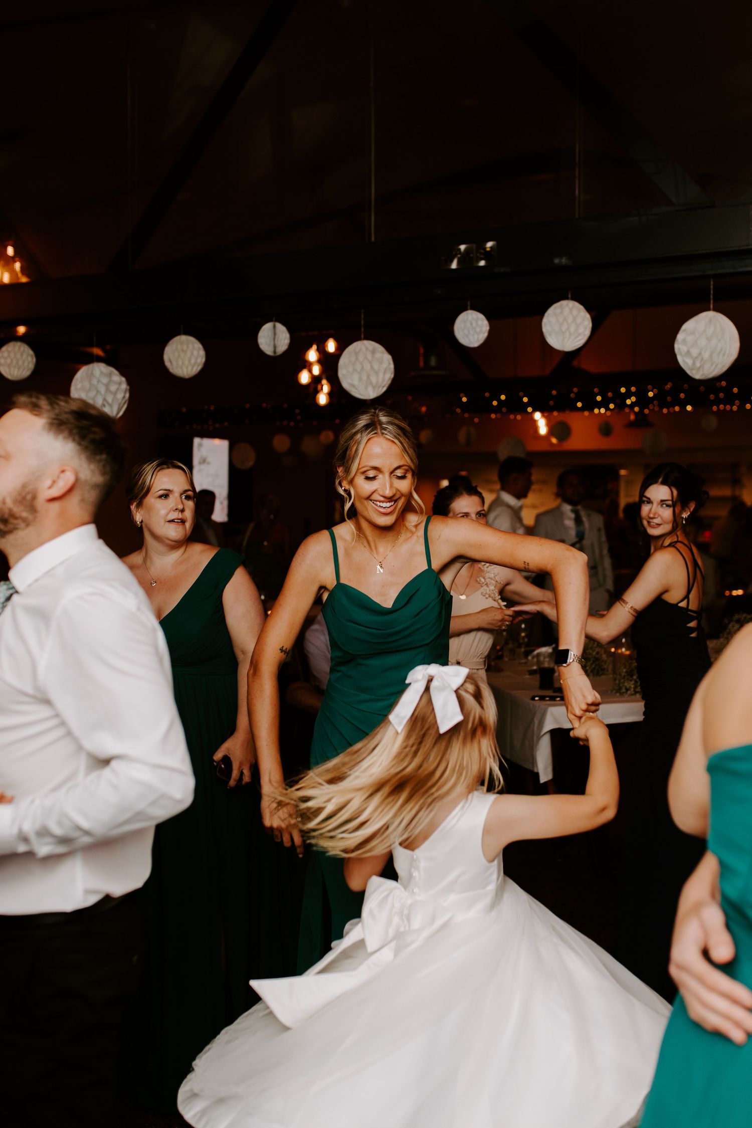 Wedding guests dance and celebrate on a dimly lit dance floor decorated with hanging white paper lanterns.
