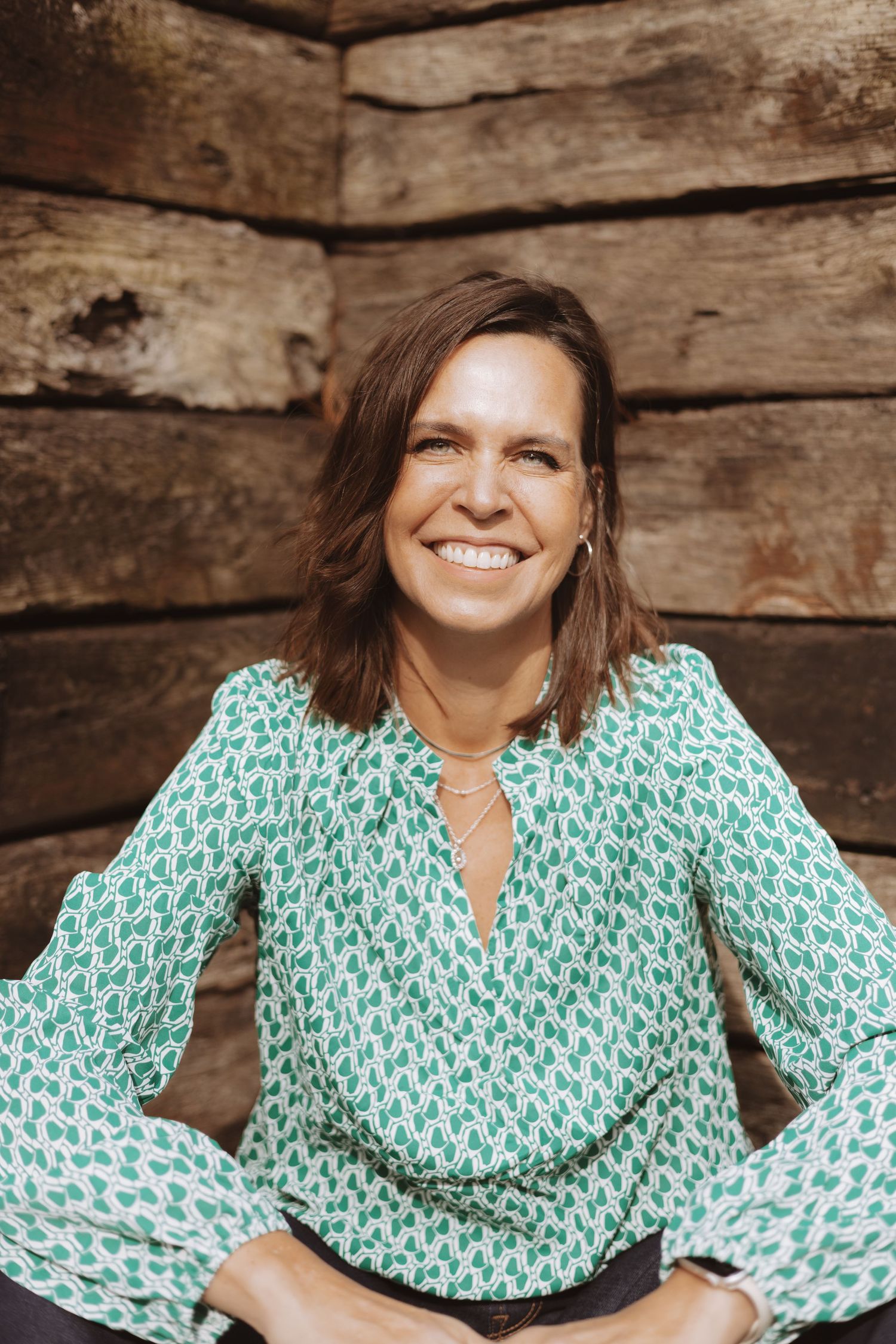 A person wearing a green and white patterned top sits against a rustic wooden wall backdrop.