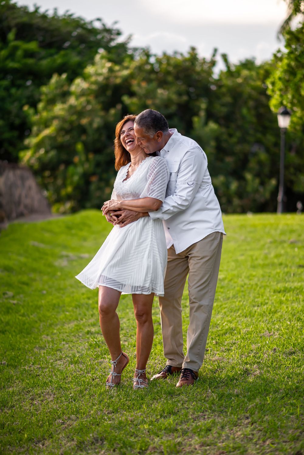 Playful couple in white outfits share tender moment on grassy hillside.