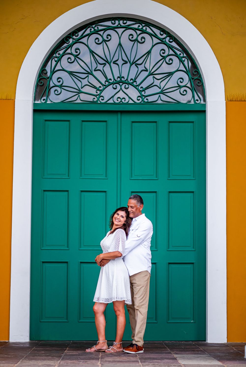 Couple poses lovingly in front of ornate turquoise colonial door with yellow walls.
