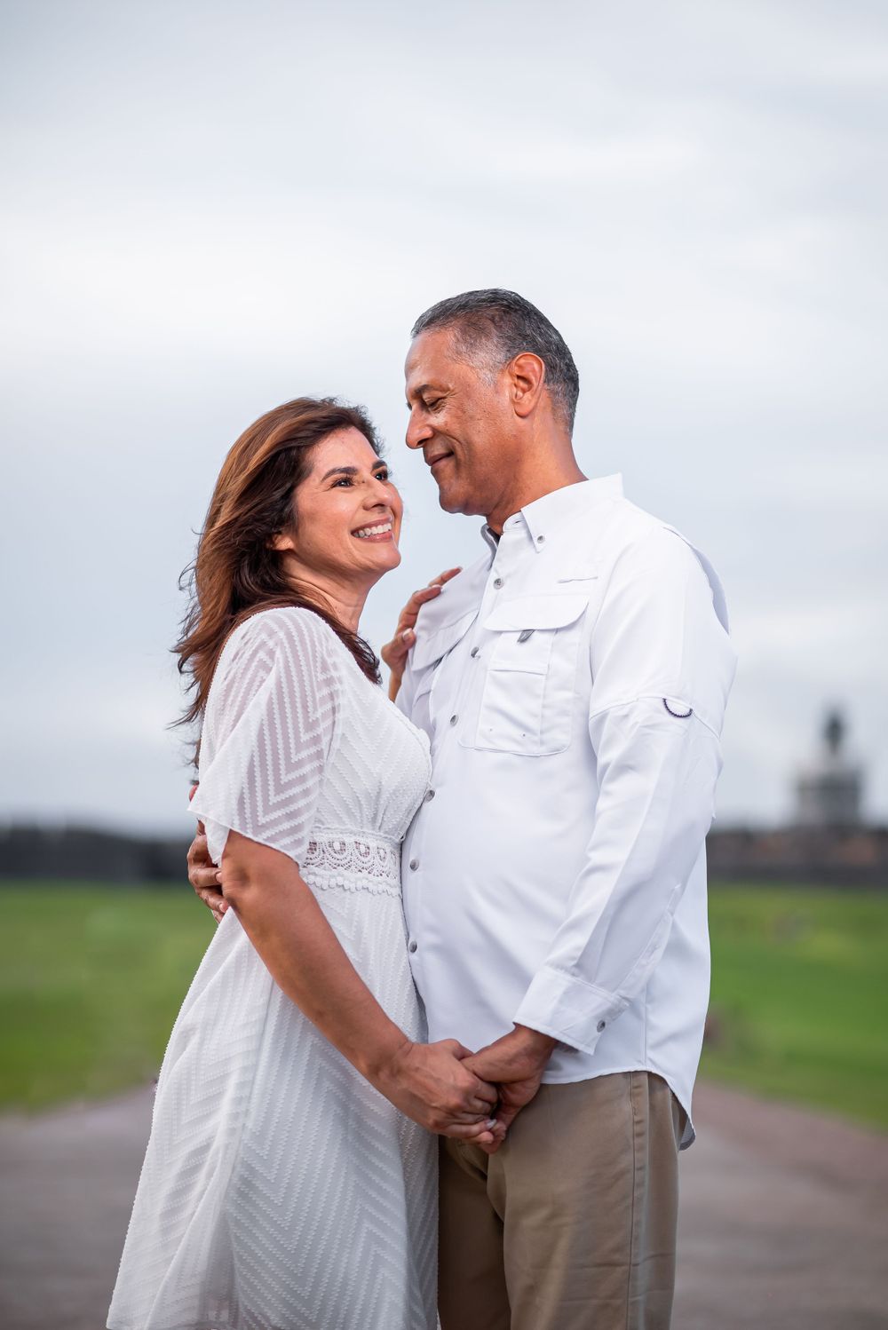 Couple in white outfits sharing intimate moment against cloudy sky.