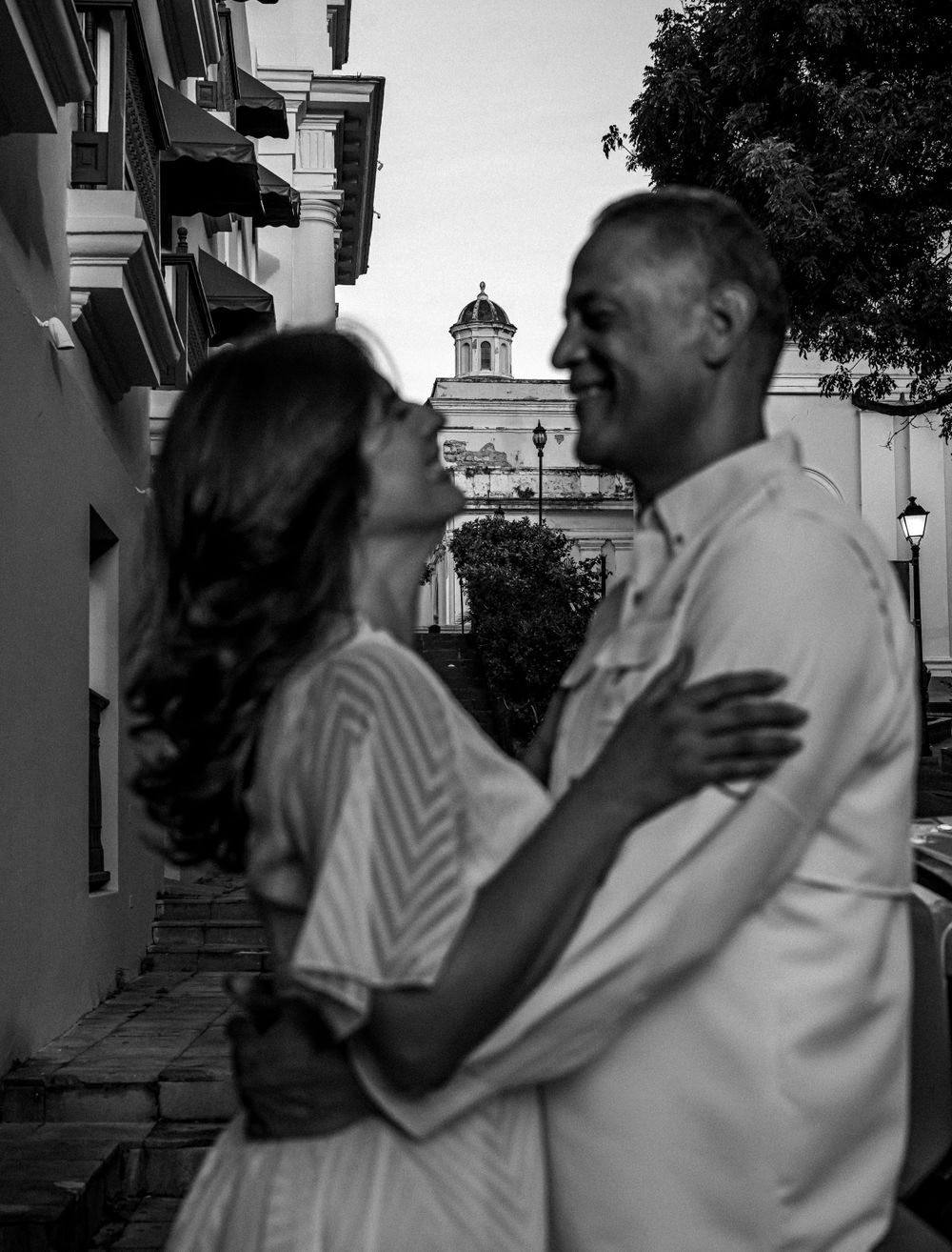 Black and white portrait of couple embracing with colonial church dome in background.