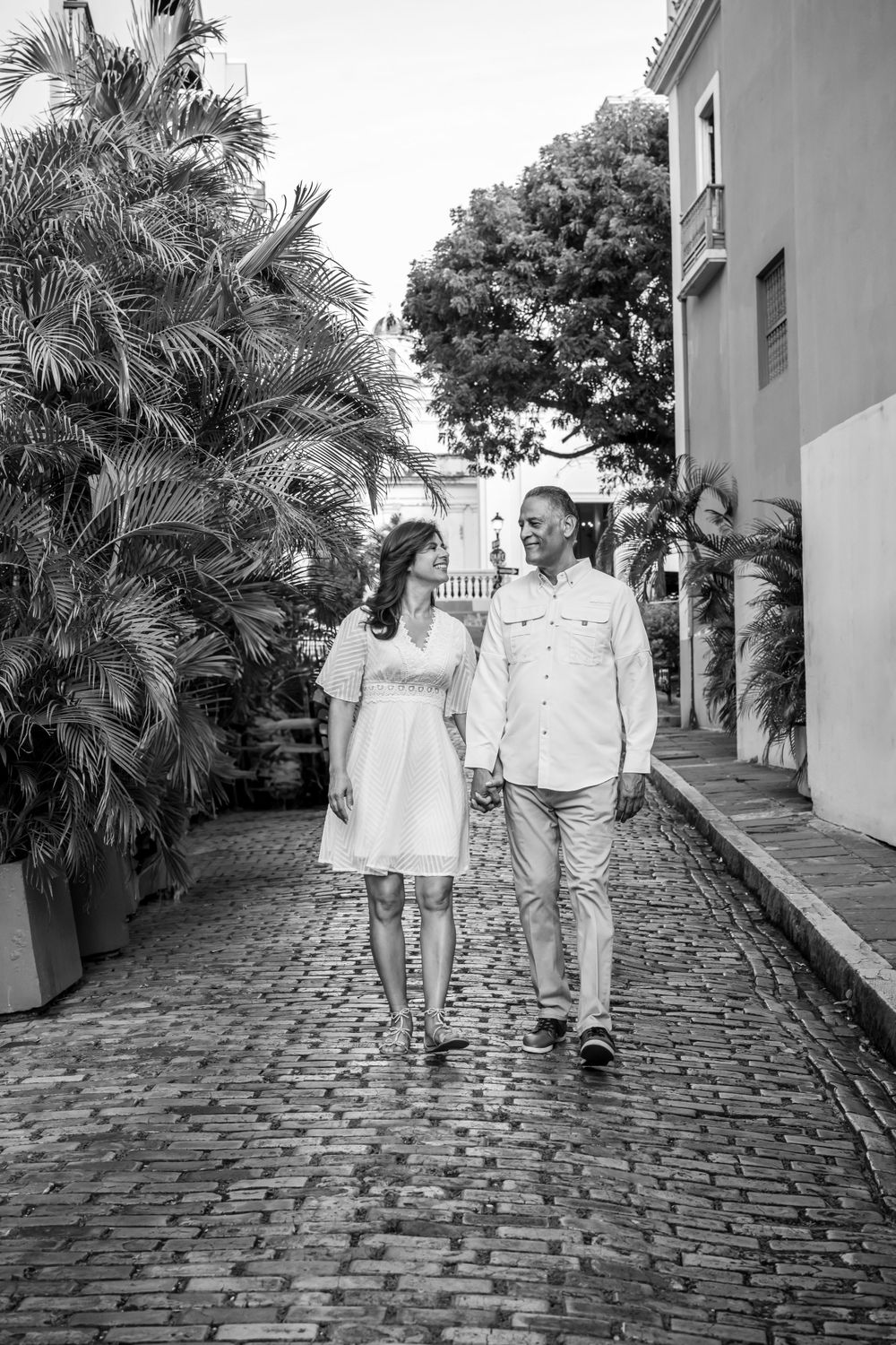 Couple in white outfits walking together on cobblestone path lined with palm trees.