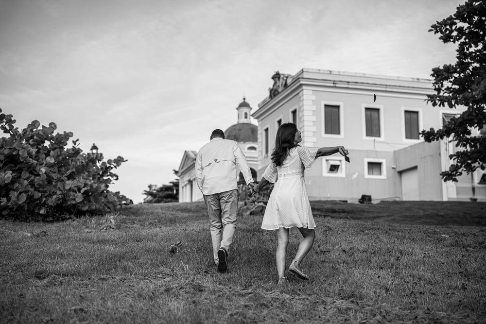 Couple holding hands while running on grassy hill with historic building in background.