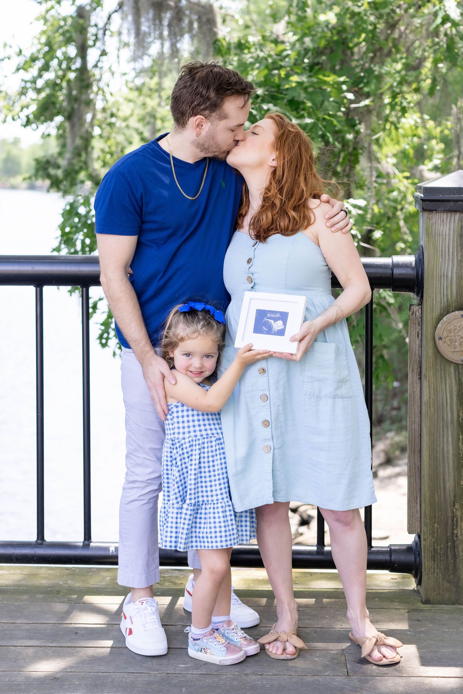 Couple kissing while daughter holding ultrasound print in outdoor maternity photo near Lake Erie, captured by Tasha Cannaday.