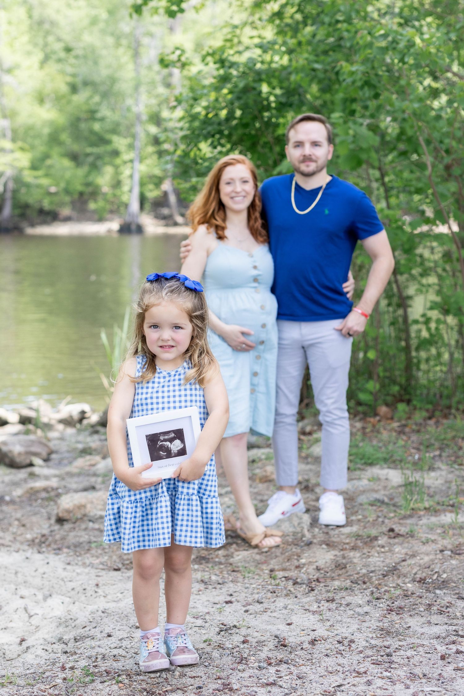 Couple smiling while daughter holding ultrasound print in outdoor maternity photo near Lake Erie, captured by Tasha Cannaday.