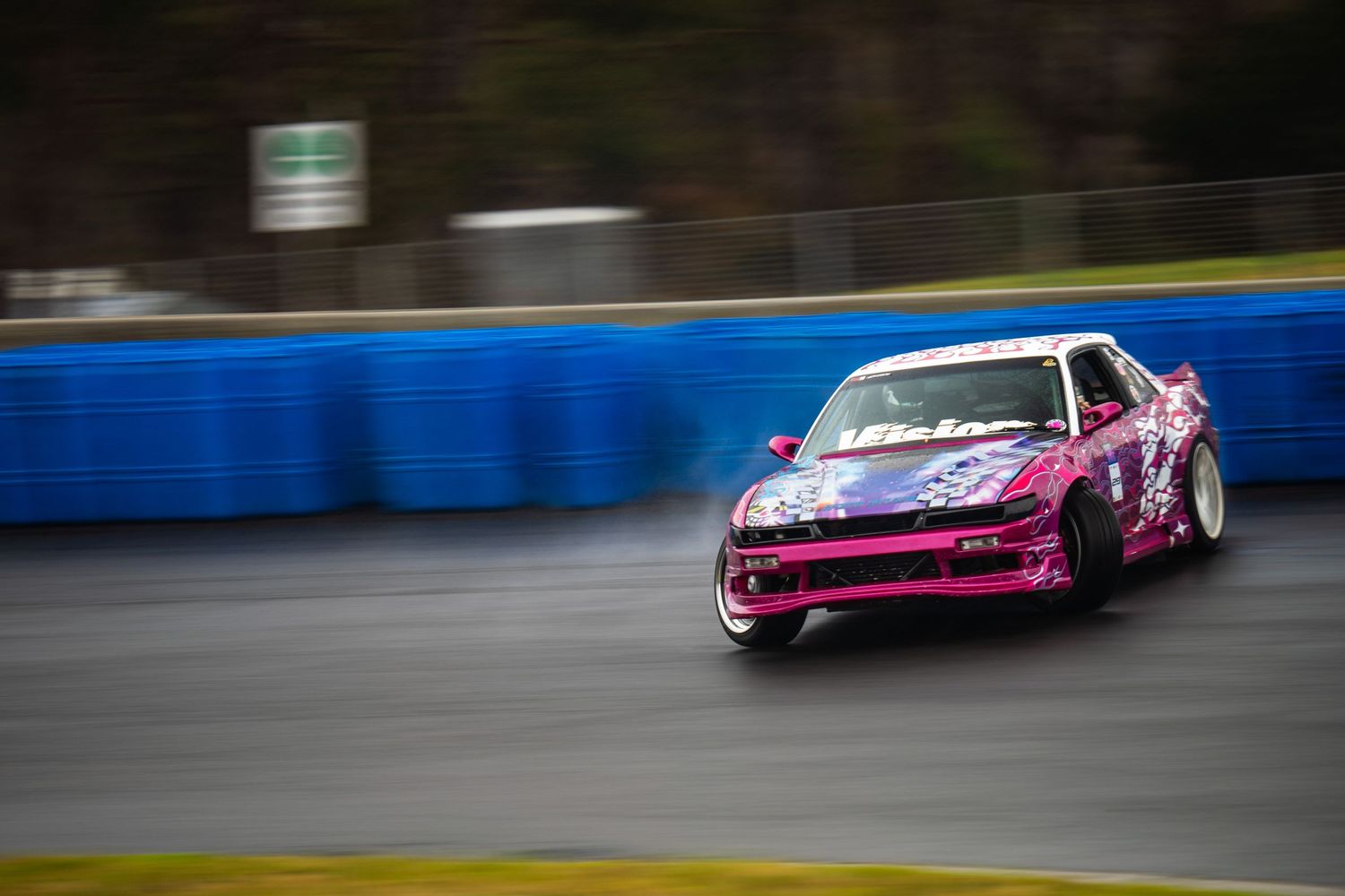 Pink drift car slides through corner on wet racing track with blue barriers.