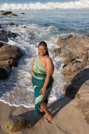 A person in a green outfit poses along rocky coastal waters as waves crash against the shore.