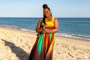 A person in a vibrant yellow and rainbow dress poses joyfully on a sunny beach with ocean waves in the background.