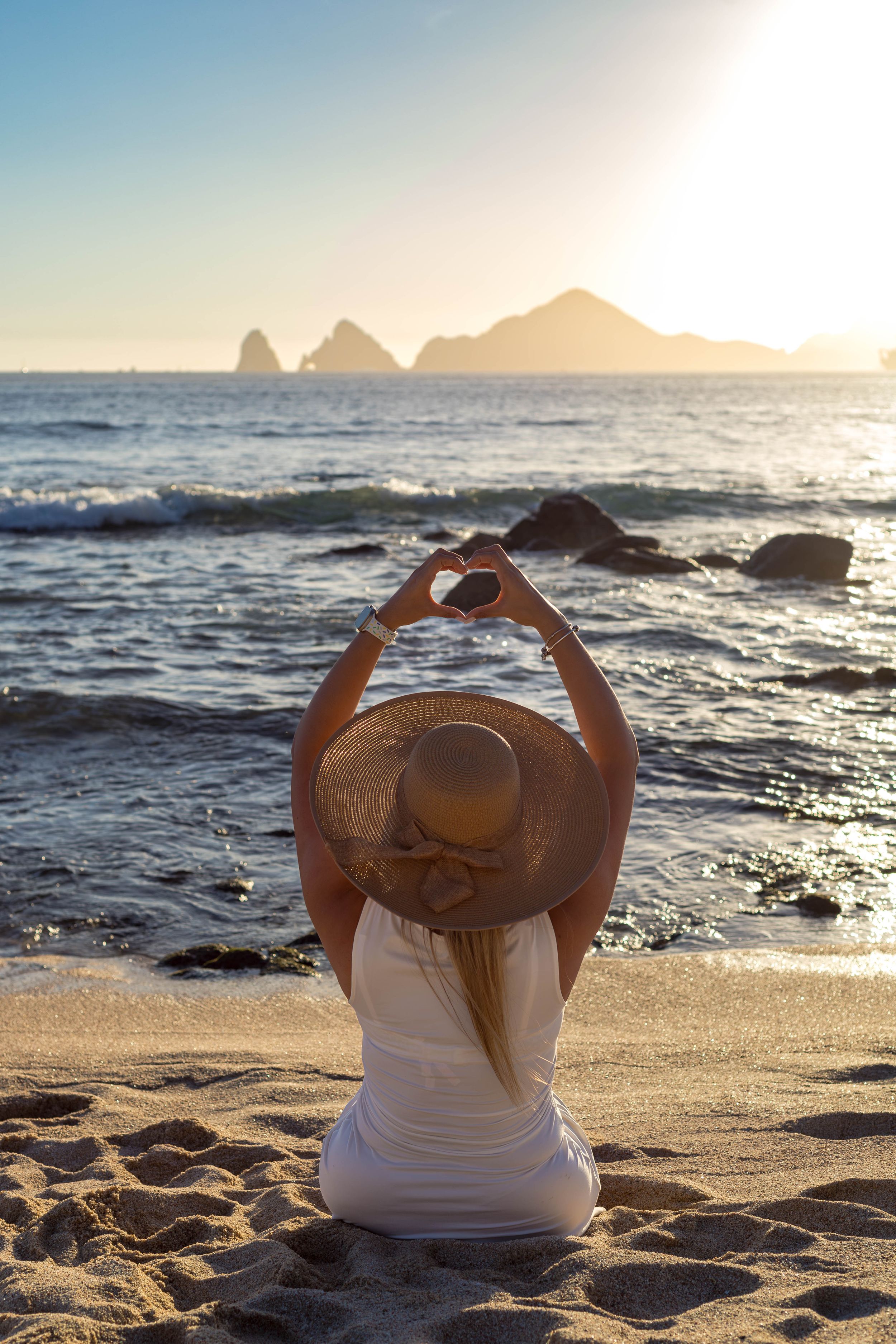 Person in white top and sun hat making heart shape with hands on beach at sunset with rocky coastline in background.