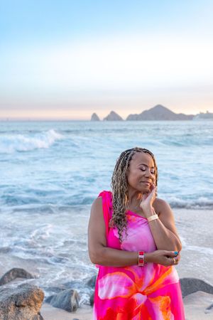 A person in a vibrant pink dress poses on coastal rocks at sunset while ocean waves crash in the background.