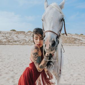 A white horse and person in a red dress pose on a sandy beach during a photoshoot.