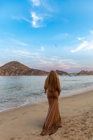 Figure in flowing mauve dress stands on sandy beach with mountain views and turquoise waters of Cabo San Lucas.