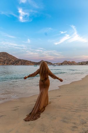 A person in a flowing beige dress stands with outstretched arms on a scenic beach with rocky mountains and blue waters.