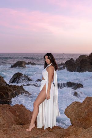 A flowing white dress contrasts against dramatic rocky coastline at sunset.