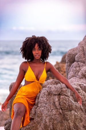 A model in a vibrant orange swimsuit poses gracefully against coastal rocks as ocean waves crash in the background.