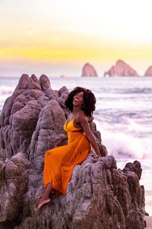 A person in a flowing orange dress poses gracefully against rocky coastal formations at sunset.