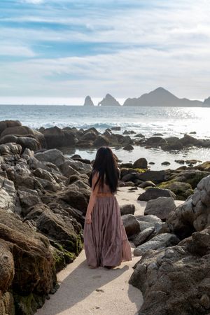 Two people sit on coastal rocks watching ocean waves crash against the rugged shoreline at sunset.