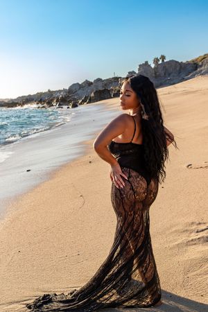 A person in a black lace dress poses gracefully on a sandy beach with ocean waves and cliffs in the background.