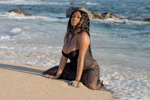 A person in a floral dress poses on a sandy beach with ocean waves in the background.