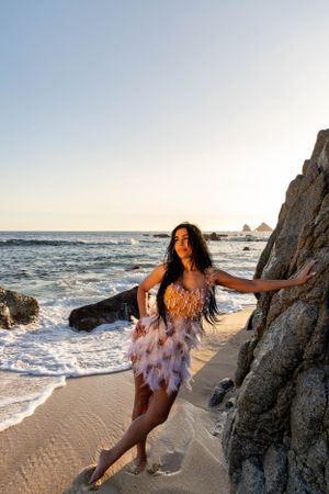 Dance-inspired beach portrait with flowing feathered dress against rocky coastline at sunset.