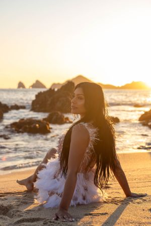 Dramatic beach portrait with long flowing hair against sunset and ocean rocks.