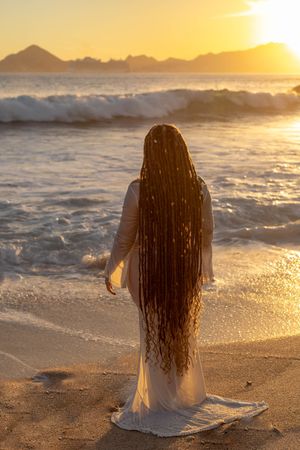 Silhouette figure with long hair standing on beach at sunset with waves crashing and mountains in background.