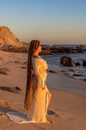 Figure in flowing yellow dress with long braids stands on sandy beach at sunset with rocky coastline in background.
