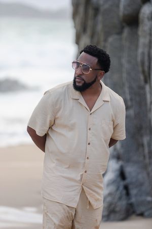 A person in a cream-colored button-up shirt poses on a beach with dark cliffs in the background.