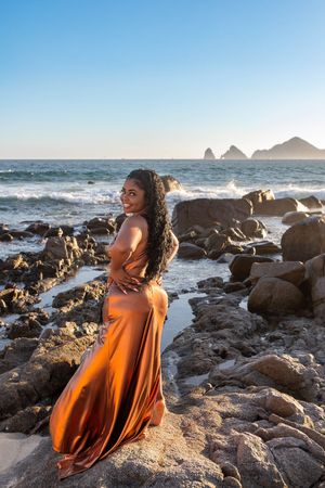Woman in flowing orange dress poses on rocky beach with ocean waves and mountain silhouettes at sunset in Cabo San Lucas.