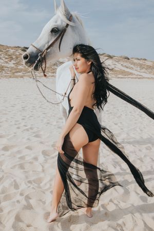A white horse stands with someone in a flowing black dress on a sandy beach landscape.