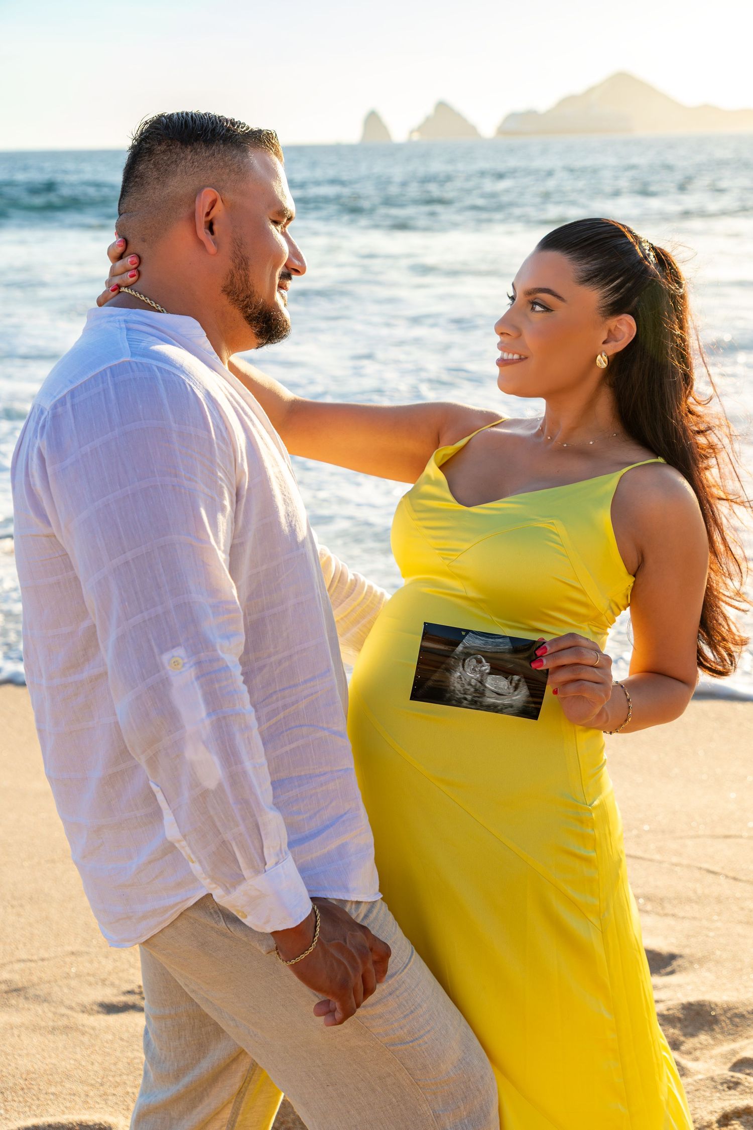 A couple in yellow and white outfits share romantic moments on a sunny beach at sunset.