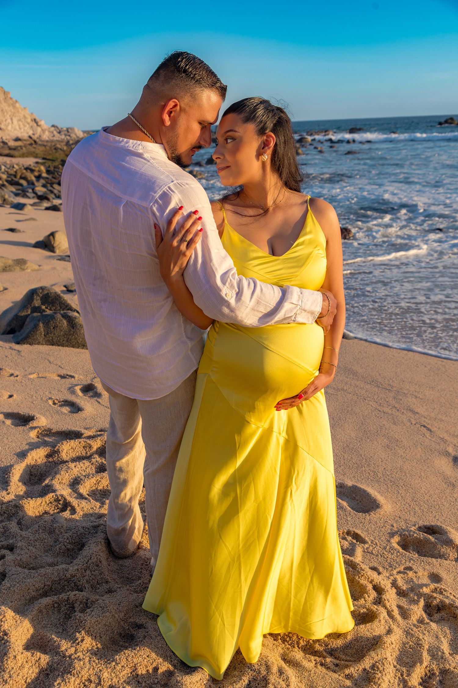 A couple embraces on a sunny beach with waves crashing behind them, the woman wearing a flowing yellow dress.