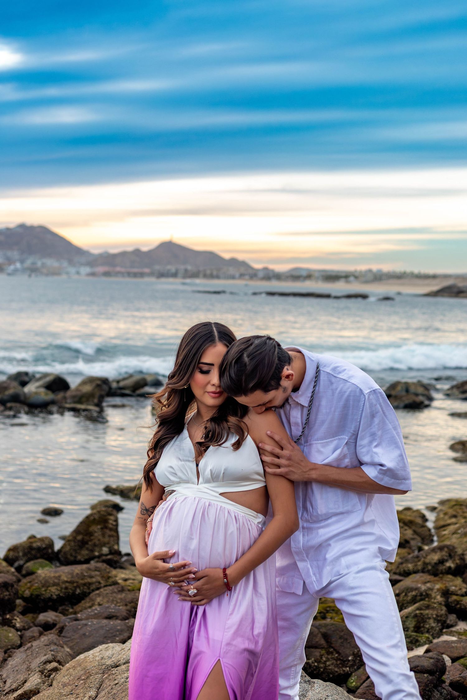 A romantic maternity photoshoot at sunset on a rocky beach with mountains and ocean waves in the background.