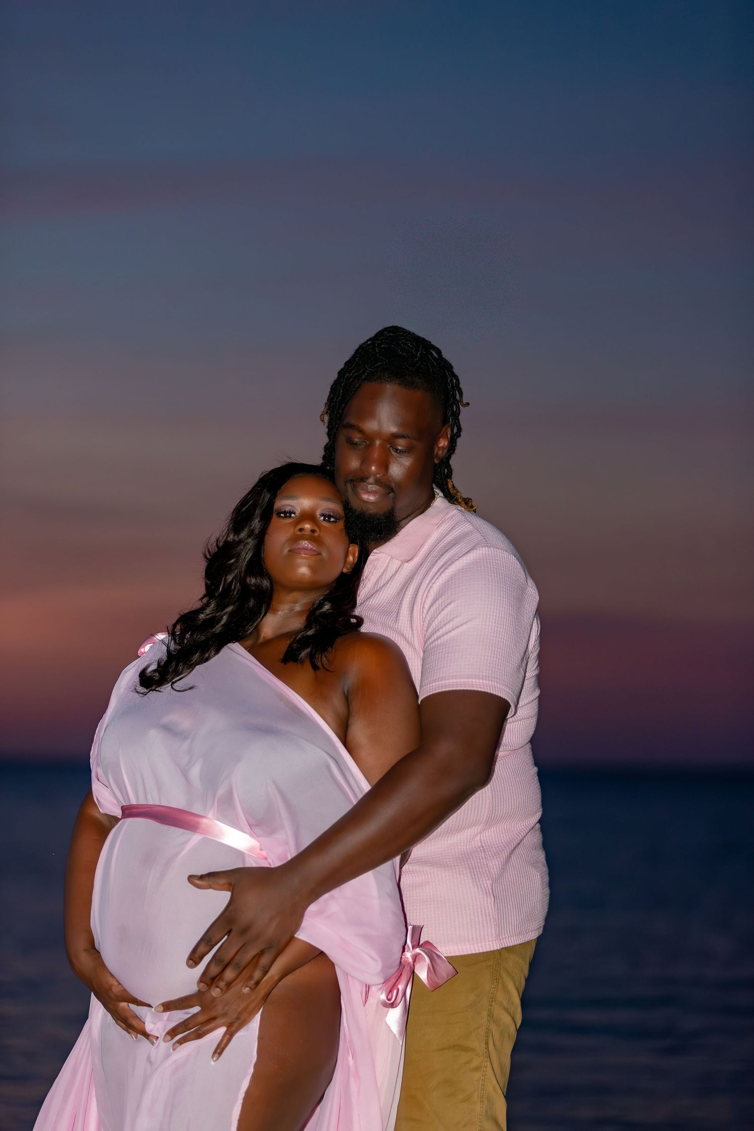 A couple in pink shirts embrace during sunset at the beach, creating a romantic silhouette against the dusky sky.