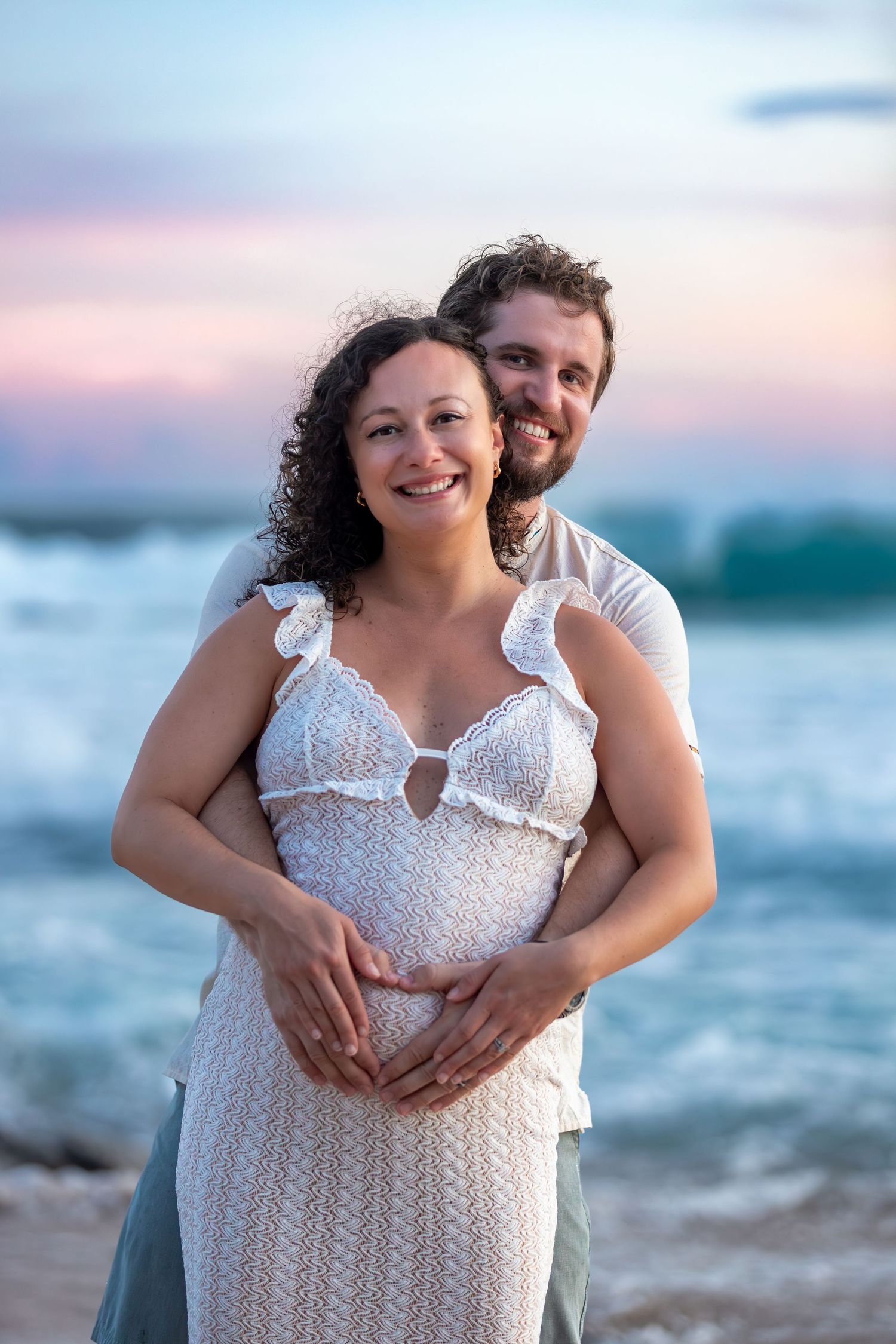 A couple embraces on a beach at sunset wearing white outfits against a pink and blue sky.