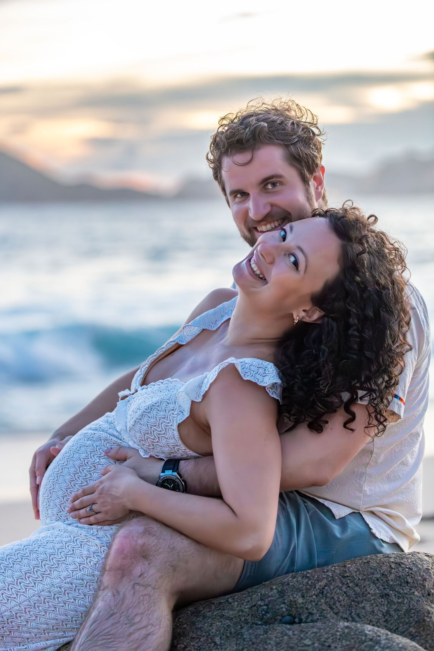 A couple embraces on a rocky beach at sunset with the ocean waves in the background.