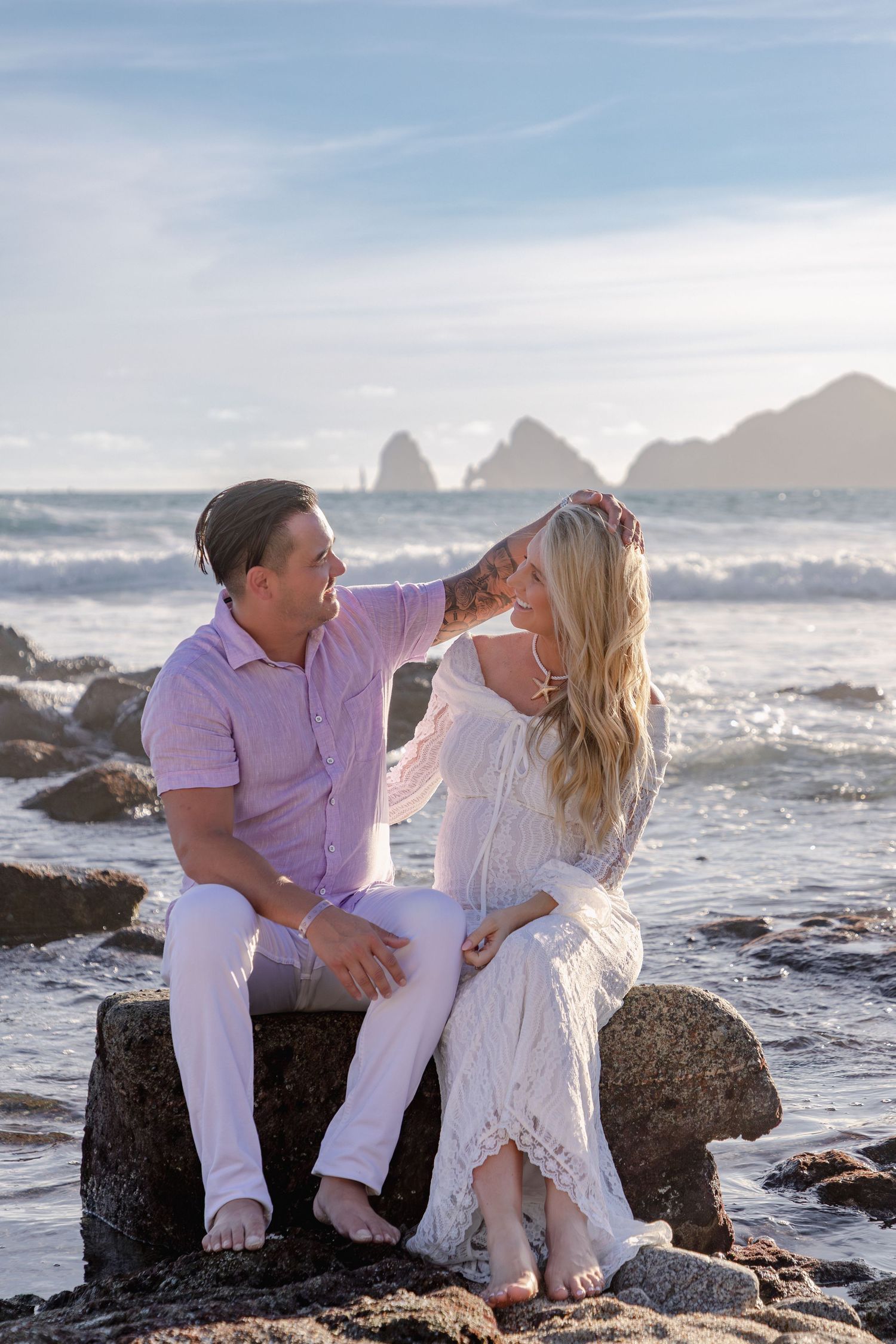 A couple in white outfits shares romantic moments on rocky coastal beach at sunset with ocean waves and mountains in background.