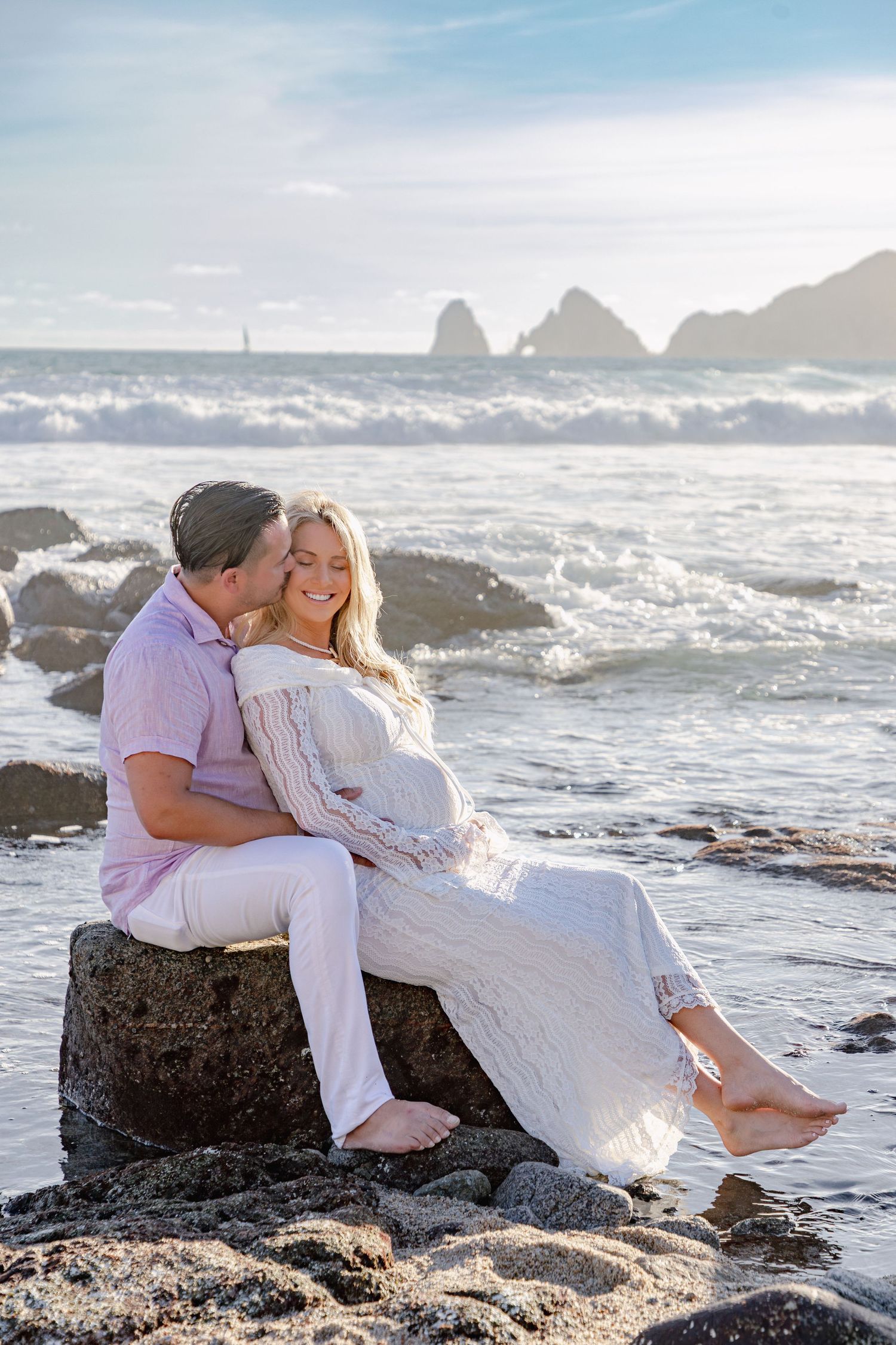 A couple shares romantic moments on coastal rocks as ocean waves crash behind them at sunset.
