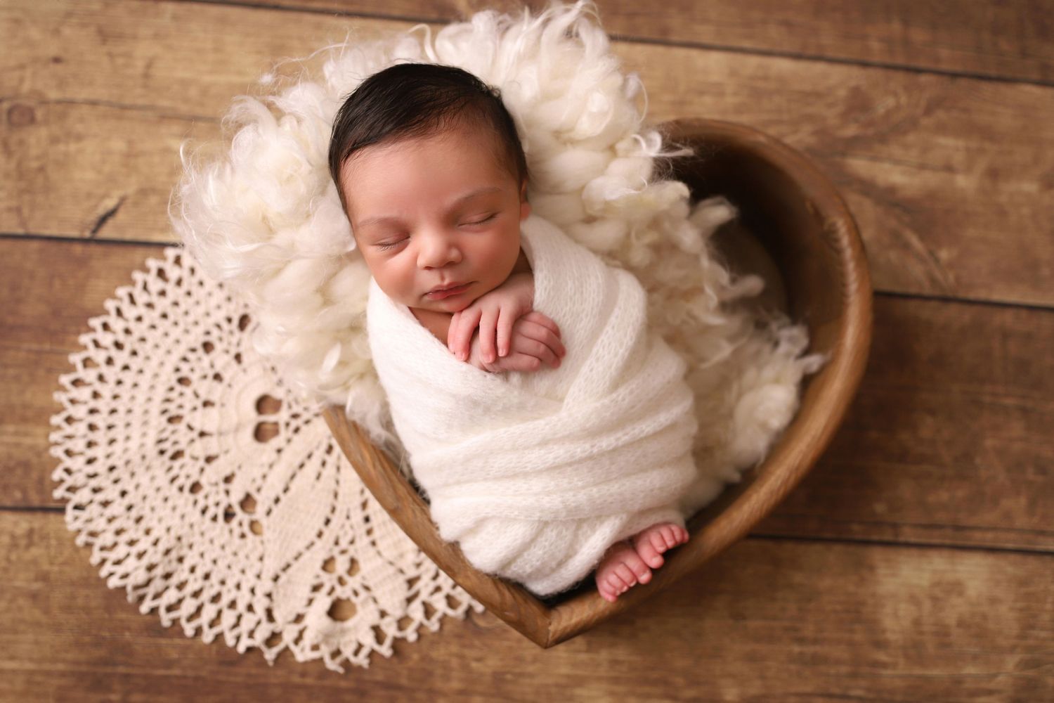 Sleeping newborn wrapped in white swaddle on lace doily against wooden background.