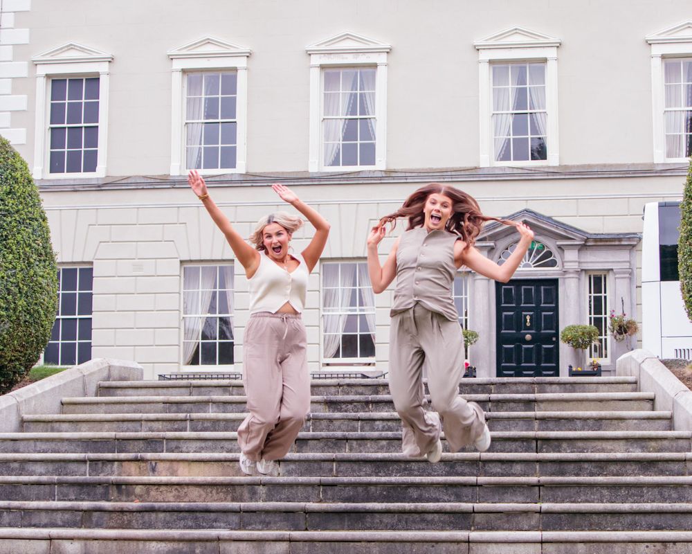 People jumping joyfully on the steps of a grand white building with multiple windows.
