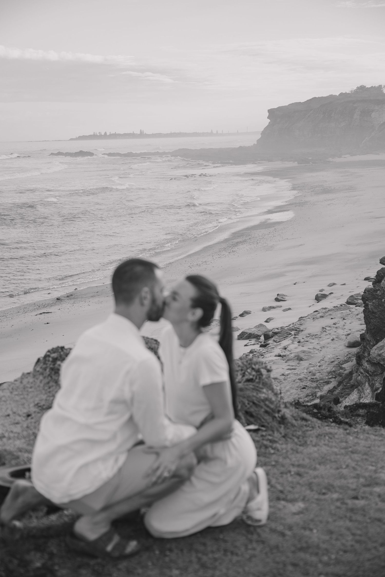 Black and white photo of a couple kissing on a grassy coastal headland overlooking a quiet beach and cliffs.