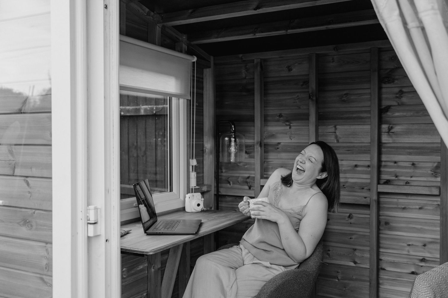 a lady is sat in her garden summer house. she is holding a cup of tea and throwing her head back and laughing with her eyes closed.