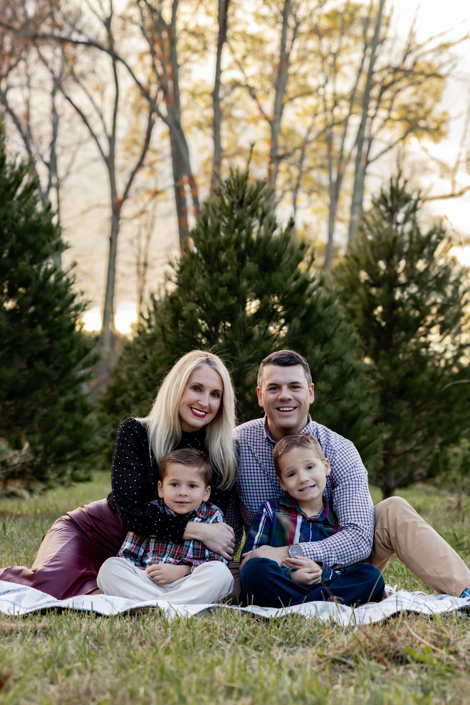 A family sits together on a blanket outdoors with evergreen trees and autumn foliage in the background. RVA Photographer family fall session